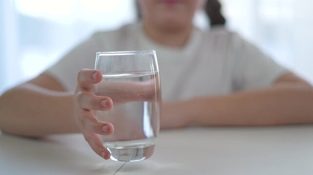 Glass filled with water close to child. Girl maintains hydration with simple water intake. Hand reaches for clean water to support child health. Water helps girl stay strong through hydration