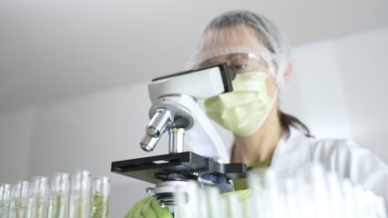 Scientist analyzing test tubes, wearing green protective gear while working with microscope in...
