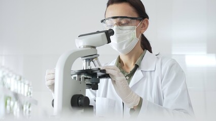 Female scientist wearing a lab coat, face mask, and safety glasses carefully adjusts a microscope,...