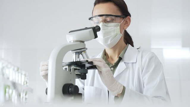 Female scientist wearing a lab coat, face mask, and safety glasses carefully adjusts a microscope, conducting research in a brightly lit laboratory setting. Medicine, healthcare and science concept