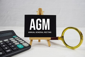 AGM abbreviation of Annual General Meeting text with magnifying glass and calculator on wooden background