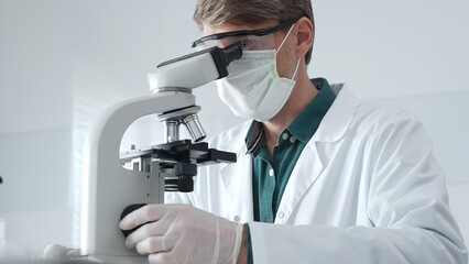 Adult male scientist wearing white mask, lab coat and protective glasses using microscope, doing...