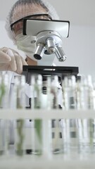 Scientist wearing green protective gear using microscope, analyzing test tubes in laboratory,...