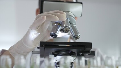 Female scientist wearing white protective gear analyzing test samples under microscope in contemporary research environment, close up of white gloved hand. Medicine and science concept