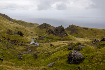 Fototapeta premium A scenic landscape with rolling green hills, rocky outcrops, and small pools of water. An overcast sky and a distant body of water add depth to this untouched natural setting.