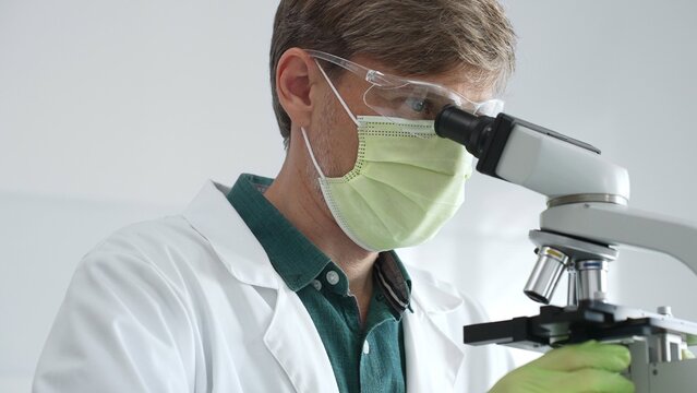 Masked adult scientist analyzing microscope slides, wearing green protective gear while conducting detailed medical research in sterile laboratory environment. Medicine, healthcare and science concept
