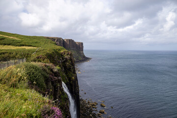 A scenic coastal landscape featuring high cliffs covered in green vegetation and a waterfall cascading into the ocean. A fence lines the edge of the cliff, and the partly cloudy sky