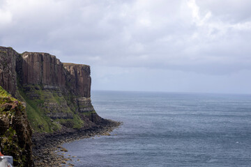 A scenic coastal landscape featuring high cliffs covered in green vegetation and a waterfall cascading into the ocean. A fence lines the edge of the cliff, and the partly cloudy sky