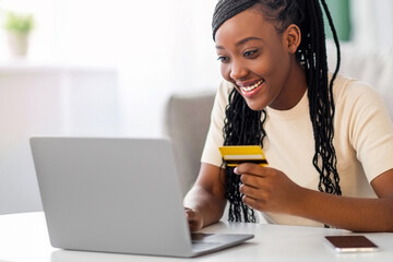 Positive young black woman holding credit card, using laptop at home, copy space. Happy african...