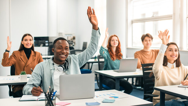 Training Class And Knowledge Concept. Diverse Group Of Smiling Multicultural People Raising Hands Up Asking Coach a Question Or Answering, Sitting With Laptops At Desks In Modern Classroom