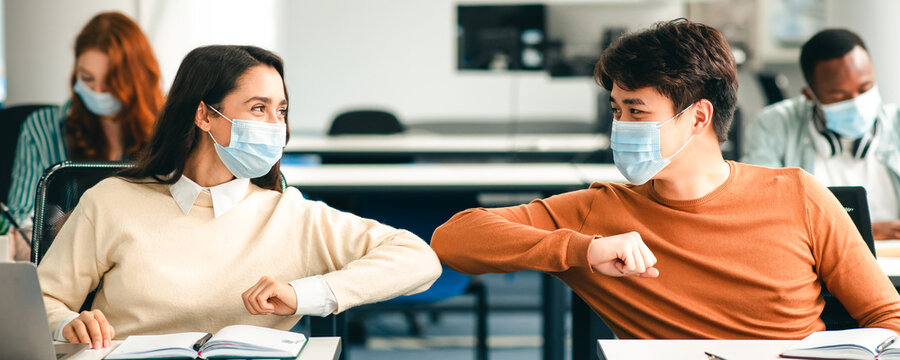 Elbow Bump. Young multiethnic students wearing protective disposable surgical masks greeting each other at classroom. Man and woman avoid touching, studying in high school, sitting at table