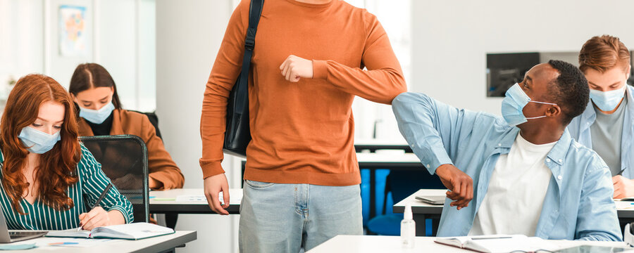 Elbow Bump. Young multiethnic students wearing protective disposable surgical masks greeting each other at classroom. Black and asian guys avoid touching, studying in highschool, college or university