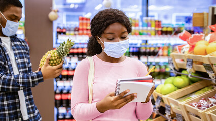 Shopping during covid-19 outbreak. Young black lady in face mask and her boyfriend buying fruits, checking grocery list at mall. Millennial couple purchasing food products at supermarket