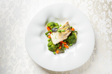Gourmet dish with cooked fish, flower garnish, and steamed vegetables including broccoli, carrots, and peas on elegant white plate.