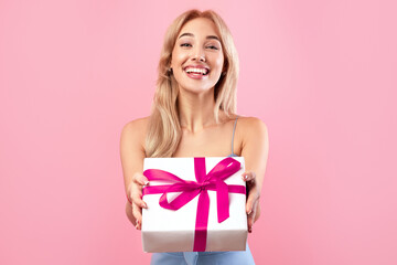 Holiday surprise. Beautiful young woman giving gift box at camera on orange studio background....