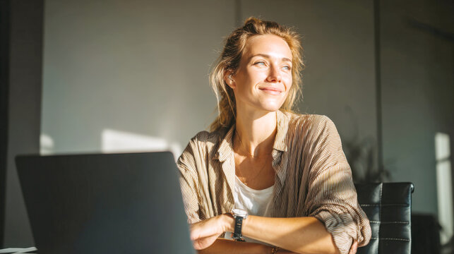 A smiling young woman sits at her home office desk with laptop, bathed in warm sunlight. This image captures essence of successful freelancer or remote worker enjoying her career and work-life balance