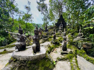 A wide-angle, medium-shot view captures a mystical garden with ancient stone statues of Thai...