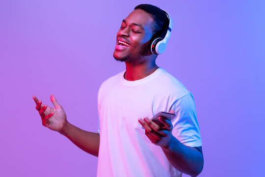Cheerful African American Guy With Smartphone Listening Music In Wireless Headphones Under Neon Lighting, Positive Young Black Man Singing With Closed Eyes, Standing Over Purple Studio Background