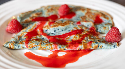 Blue pancakes are beautifully arranged on a white plate, topped with fresh raspberries and raspberry sauce, accompanied by a cup of coffee on a gray surface.