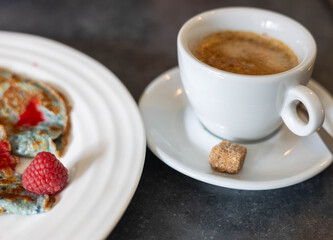 A delicious breakfast spread features colorful blueberry pancakes topped with fresh raspberries, paired with a warm cup of coffee and a sugar cube, all set on a dark table.