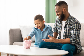 Personal Savings. African Father And Son Putting Coin In Piggybank Sitting On Sofa At Home, Dad Teaching His Kid Financial Literacy, Talking About Money And Budget Planning. Selective Focus