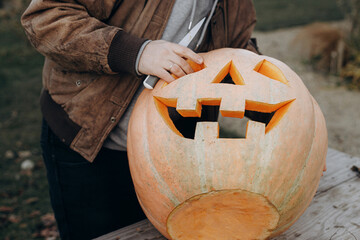 Stylish man carving halloween pumpkin on rustic wooden table in autumn garden. Fall harvest....