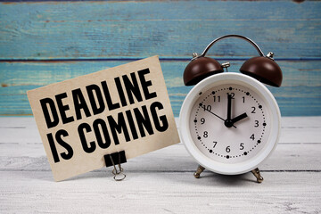 Deadline is Coming text written on paper card with alarm clock on wooden background