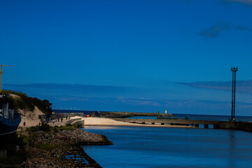Obraz premium Scenic view of a sandy beach promenade, pier, and green lighthouse under a deep blue sky with calm water and visitors.