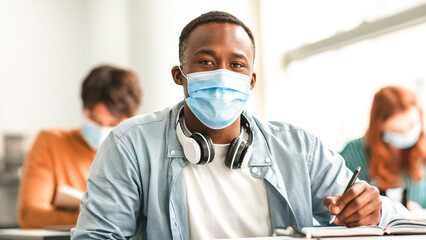 Education During Pandemic Concept. Portrait of black male student sitting at table in classroom at university, wearing protective medical face mask, writing in notebook, looking posing at camera