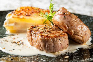 Gourmet lasagna with golden crust served alongside grilled meat medallions, garnished with fresh greens and creamy sauce on an elegant black speckled plate.