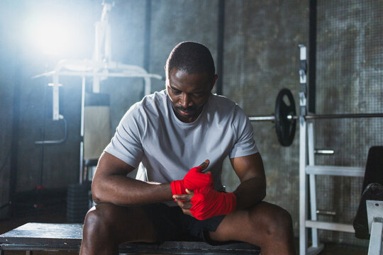Fitness workout in gym. African American man fighter preparing for fight wrapping hands with red boxing wraps sports protective bandages in gym. Strong man ready for fight boxing sparring training
