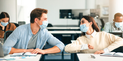 Elbow Bump. Young multiethnic classmates wearing protective disposable surgical masks greeting each other at classroom. Smiling man and woman avoid touching, studying in highschool, sitting at table