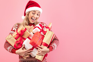 Happy Caucasian lady in cozy warm sweater and Santa hat holding heap of gifts over pink studio...