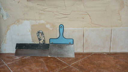 Poor-quality wall renovation with uneven plaster, visible flaws, and two putty knives on tiled...