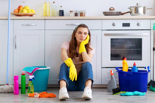 Exhausted young housewife sitting on floor at kitchen, surrounded by cleaning supplies, tired from housework. Overworked woman needing break from domestic duties, cannot stand doing house chores
