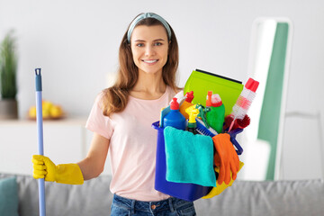 Cheerful millennial housewife holding bucket full of cleaning supplies at modern apartment....