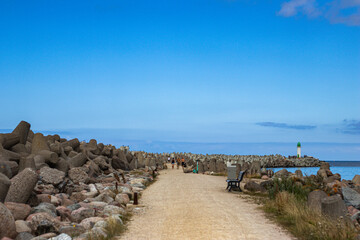 Scenic seaside path with concrete tetrapods, benches, people walking, and a green-topped lighthouse...