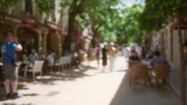Cafe patrons enjoy outdoor dining on a sunny street in mallorca with a defocused background filled with trees and blurred people walking
