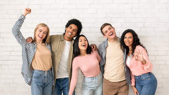 Have fun with friends and get education in college or university. Happy young multiethnic people team in casual hugging and looking at camera, on white brick wall background, studio shot