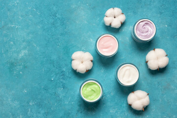 Colorful cosmetic creams in jars on blue grunge background decorated with fluffy cotton flowers, top view, flat lay, copy space, natural organic cosmetics