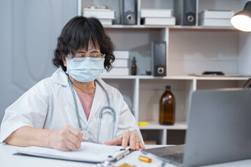 A doctor in a white coat sits at a clinic desk, writing prescriptions for patients and recording...