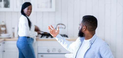 Hungry african husband complaining about empty plate, waiting for dinner in kitchen while his spouse cooking food on background, starving black man sitting at table and demanding food, free space