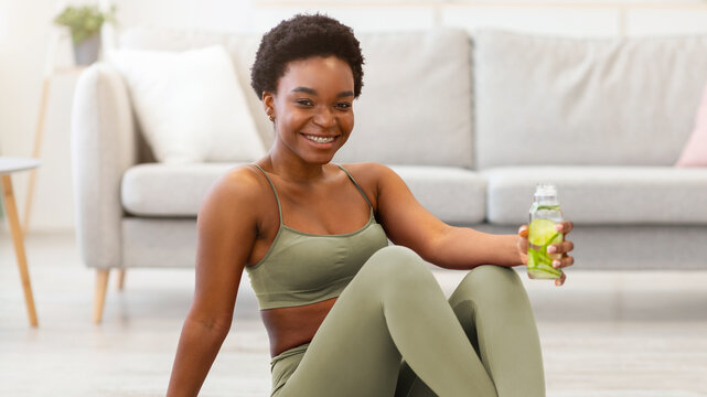 Stay Hydrated. African Fit Woman Holding Bottle Of Infused Water Drinking During Workout Sitting On Gymnastics Mat Posing At Home, Smiling To Camera. Healthy Hydration And Weight Loss Concept