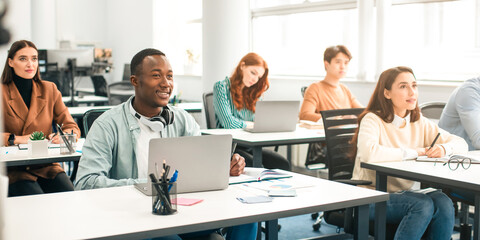Education, Technology, Lifestyle And People Concept. Group of diverse international students sitting at desk in classroom using laptop computers, studying and listening to teacher, writing in notepads