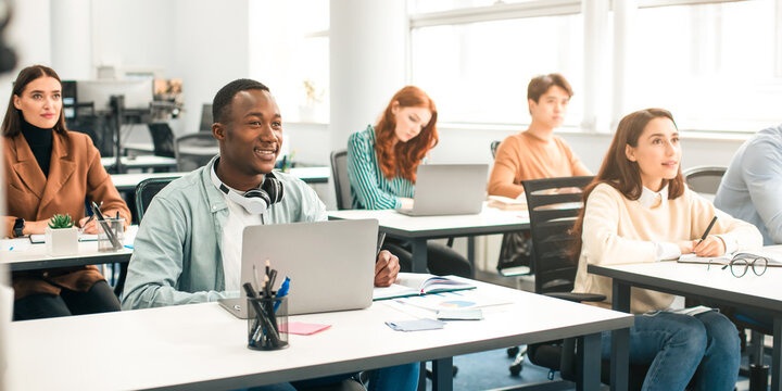 Education, Technology, Lifestyle And People Concept. Group of diverse international students sitting at desk in classroom using laptop computers, studying and listening to teacher, writing in notepads