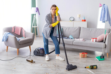 Unhappy young woman standing with vacuum cleaner in messy flat after party, blank space. Upset millennial housewife at dirty room after celebration, garbage scattered everywhere