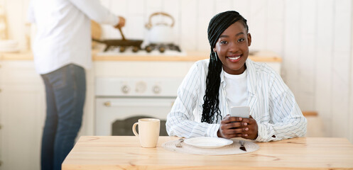 Young Black Woman Sitting At Table In Kitchen With Smartphone While Her Husband Cooking Lunch On Background, Millennial African American Couple Spending Time Together At Home, Selective Focus