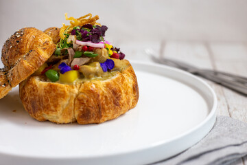 A flaky, golden croissant is filled with creamy savory filling, topped with microgreens, edible flowers, and colorful vegetables, served open-faced on a clean white plate for an elegant presentation.
