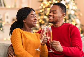 Happy family and romantic date at home. Young african american guy and woman clink glasses of champagne sit on sofa in living room interior with christmas tree with glowing garlands, free space