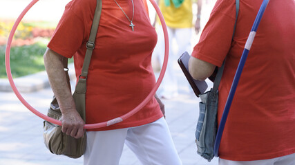 Two senior women dressed in red T-shirts and white pants stand outdoors holding hula hoops during a fitness activity session in a park, promoting healthy aging and active lifestyle for older adults.